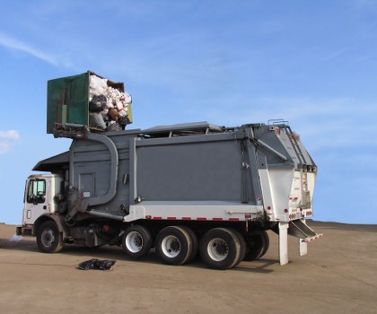 A large commercial waste collection truck parked on a flat, paved surface with a clear blue sky in the background. The truck has a white cab and a large, dark grey industrial waste container mounted on the chassis. The rear section of the container features a hydraulic lifting mechanism used for loading and unloading rubbish. In the upper front part of the container, a green skip is partially visible, filled with mixed waste materials including plastic bags, cardboard, and other refuse. The waste materials appear to be loosely packed and some are spilling over the side of the skip. The area surrounding the truck is open and free of other objects, with no visible buildings or natural features, reflecting a typical site for commercial waste collection in a location that could be near Kings Cross or in central London. The scene is well-lit, emphasizing the metallic finishes and textures of the waste container and the cluttered contents of the skip, consistent with rubbish removal operations conducted by Commercial Waste Removal Kings Cross.