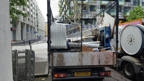 A flatbed waste collection truck parked on a city street near modern residential buildings, filled with various discarded items including broken furniture, wooden panels, and metal objects, ready for rubbish removal services. The truck features a black metal cage on the rear to secure the debris, and the vehicle has a rusted, utilitarian appearance with a yellow license plate marked 'NX12 GHA.' The surrounding environment shows a paved sidewalk and an intersection with traffic lights and street signs visible in the background, indicative of an urban area possibly within Kings Cross or nearby postcode region. The scene captures the typical process of waste collection from commercial or residential locations, managed by a company specialized in rubbish removal within the local area, with the presence of other city traffic and infrastructure elements in the background, emphasizing a professional approach to waste management services.