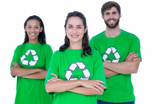 A group of three individuals standing outdoors in front of a neutral background, wearing bright green T-shirts with white recycling symbols on the front, representing a waste management or rubbish removal service. The woman in the centre has shoulder-length dark hair and is smiling with her arms crossed, while the woman on the left has long dark hair and is also smiling with her arms crossed. The man on the right has short dark hair, a beard, and is smiling with his arms crossed as well. Their attire and posture suggest a professional yet approachable team, likely from a rubbish removal company such as Commercial Waste Removal Kings Cross, serving local areas around postcode N1, with the focus on environmentally conscious waste disposal solutions in the Kings Cross district. The image emphasizes teamwork and a commitment to sustainable waste management practices, suitable for visuals on a website dedicated to commercial rubbish collection services in central London.