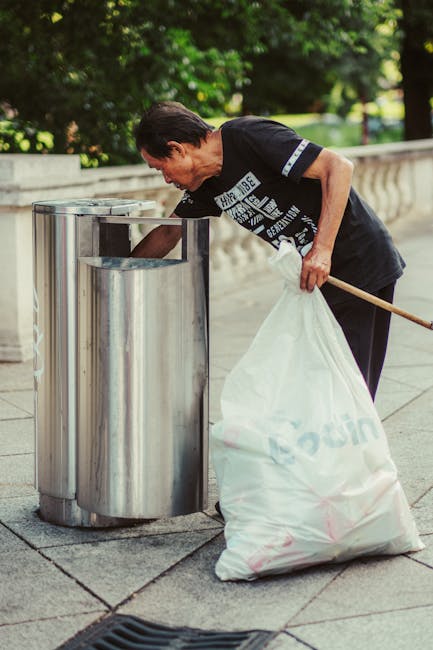 A narrow urban alleyway filled with a large pile of mixed waste, including cardboard boxes, plastic bags, and other refuse, stacked against the back wall. In the foreground, there is a wheeled black g