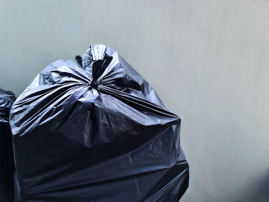 A large accumulation of mixed waste and rubbish bags, including black, white, and plastic bags along with cardboard boxes and various packaging materials, is piled on a paved area adjacent to a parkin