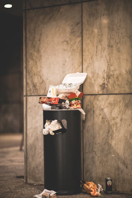 A pile of mixed waste including black plastic bin bags, some stuffed with trash, stacked on top of a yellow plastic container and an old, worn car tyre lying on its side, all situated on an asphalt su